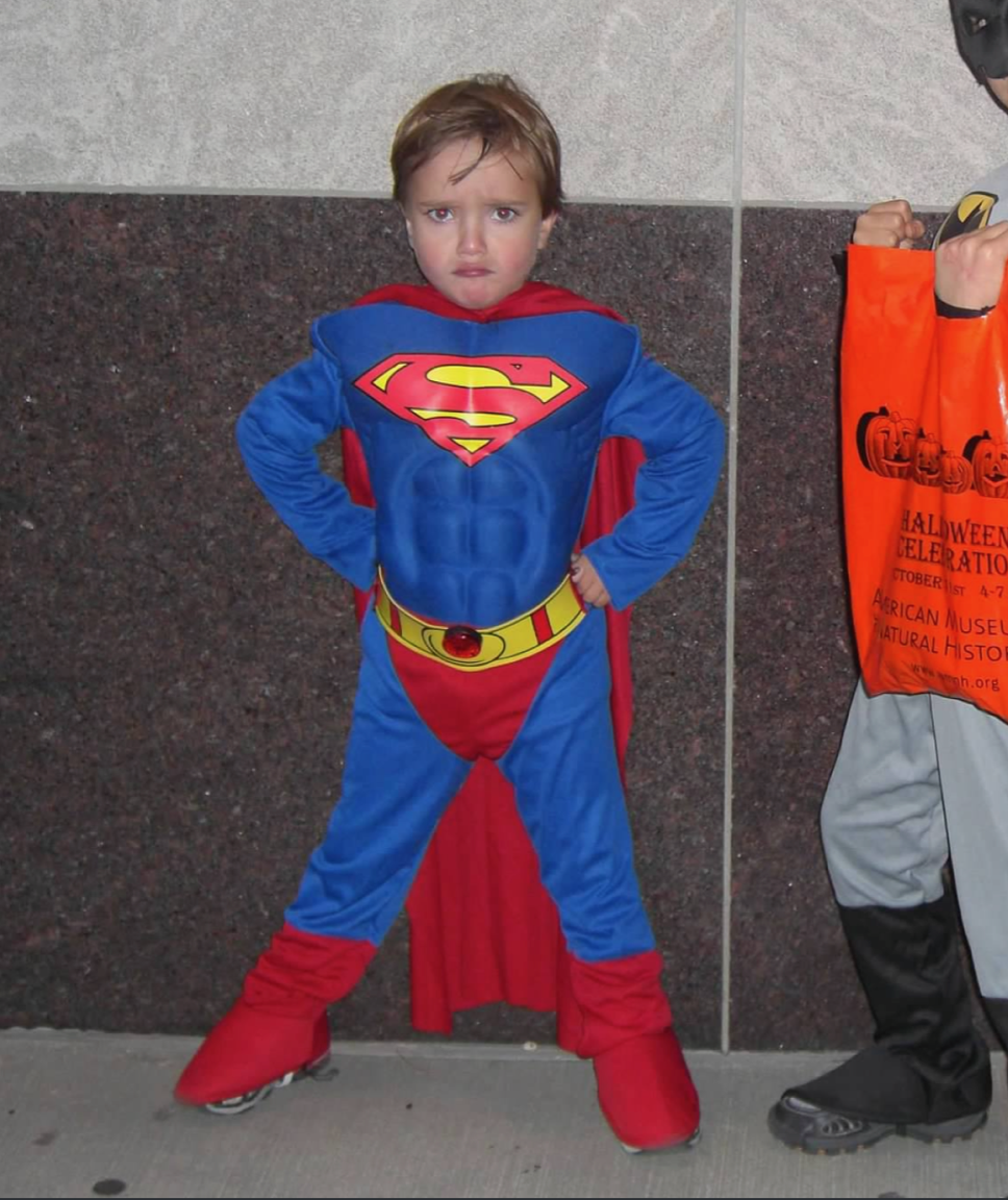 A little boy in superman costume standing next to an orange bag.